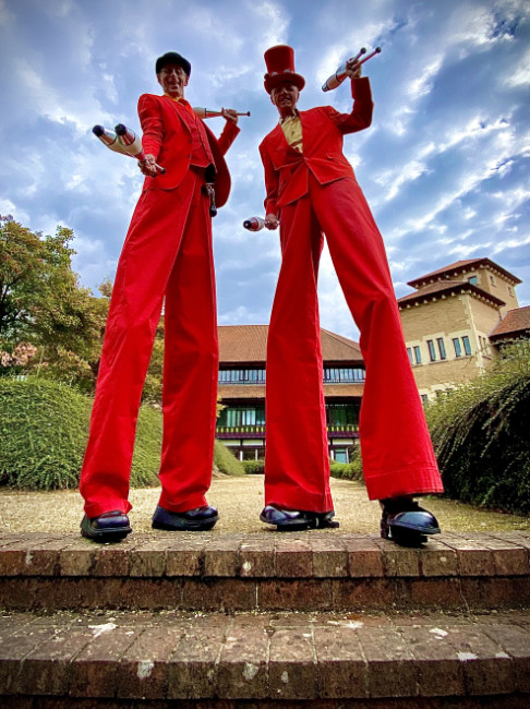 red stilts Brett Sirrell on stilts at a corporate funday in company colours and a bright red suit holding juggling clubs next to another stilt walker in the summer event